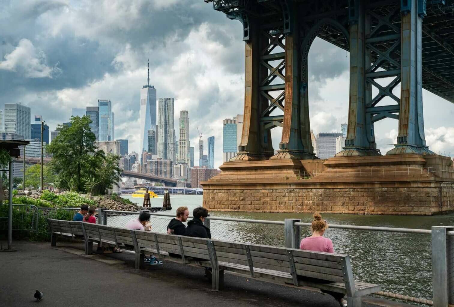 people sitting on benches under the Manhattan Bridge in Brooklyn Bridge Park in DUMBO
